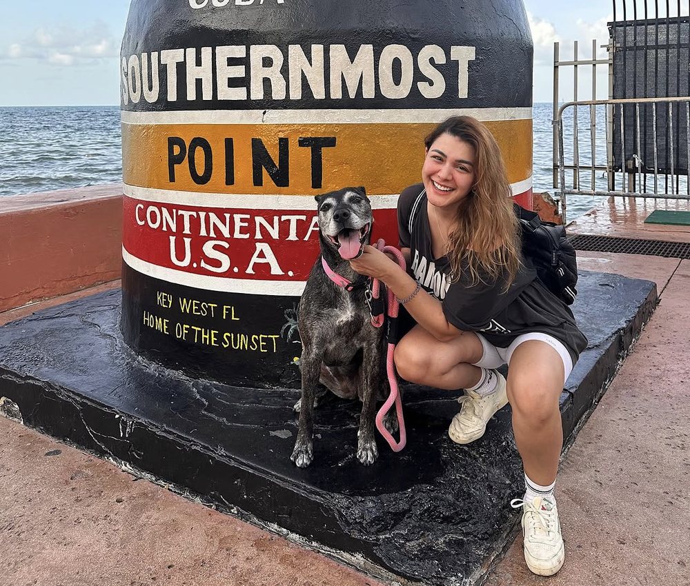Cata crouching next to Cinnamon, a happy senior brindle dog, at the Southernmost Point buoy in Key West